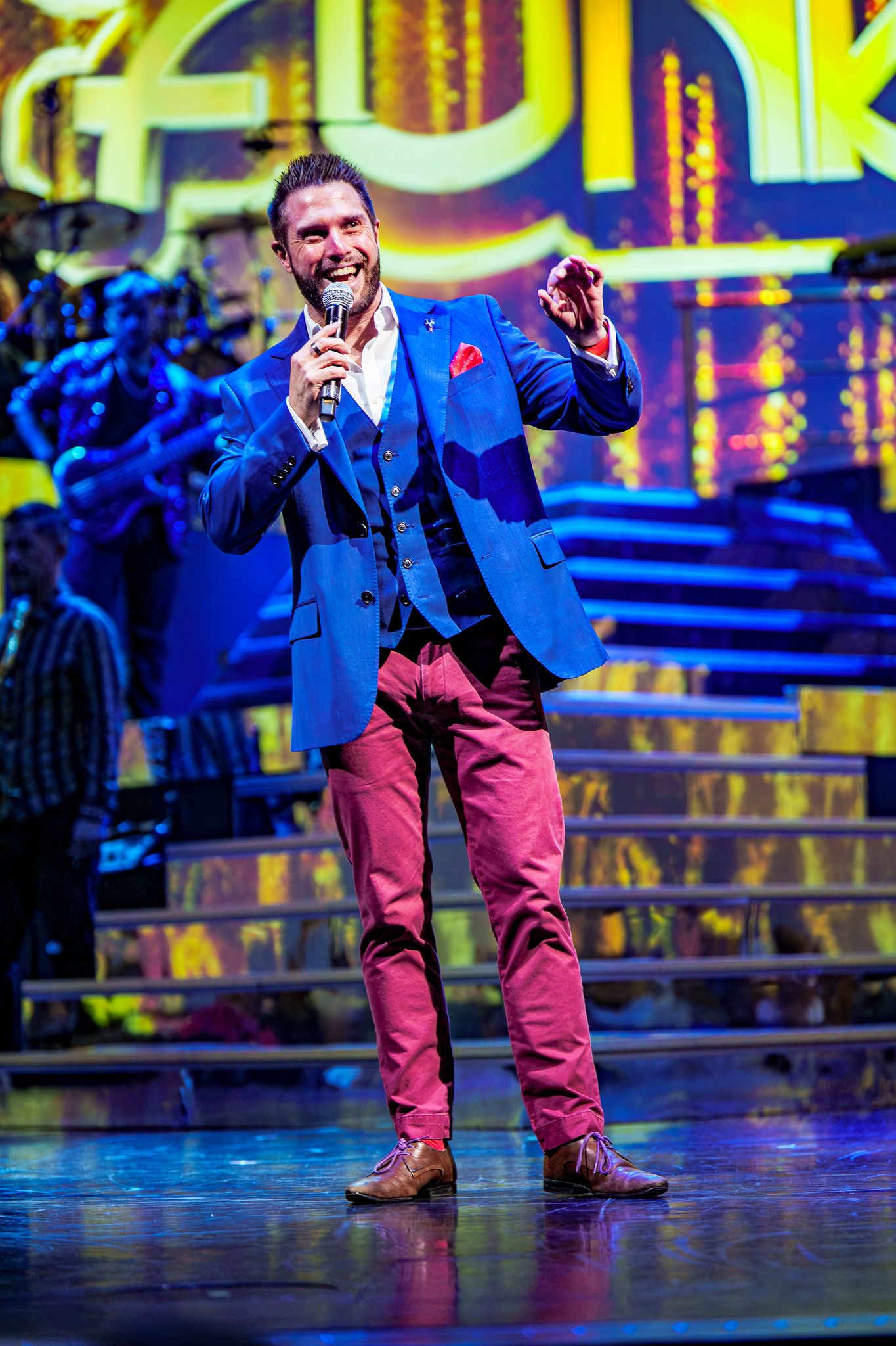 Alexander Percival hosting on stage with a microphone, wearing a blue jacket under colourful stage lights