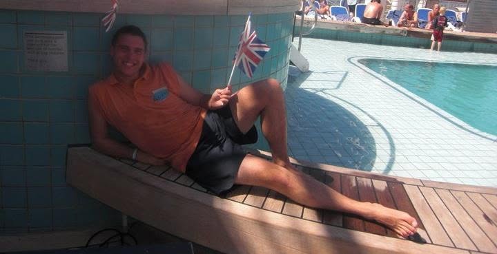 Alexander Percival in an orange Youth Staff shirt, sitting beside a ship’s pool holding a Union Jack flag